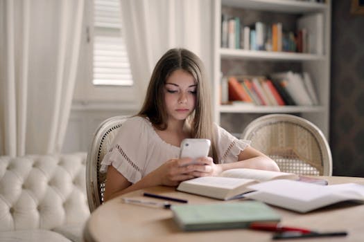 A teenage girl focused on her smartphone while studying at an indoor table.